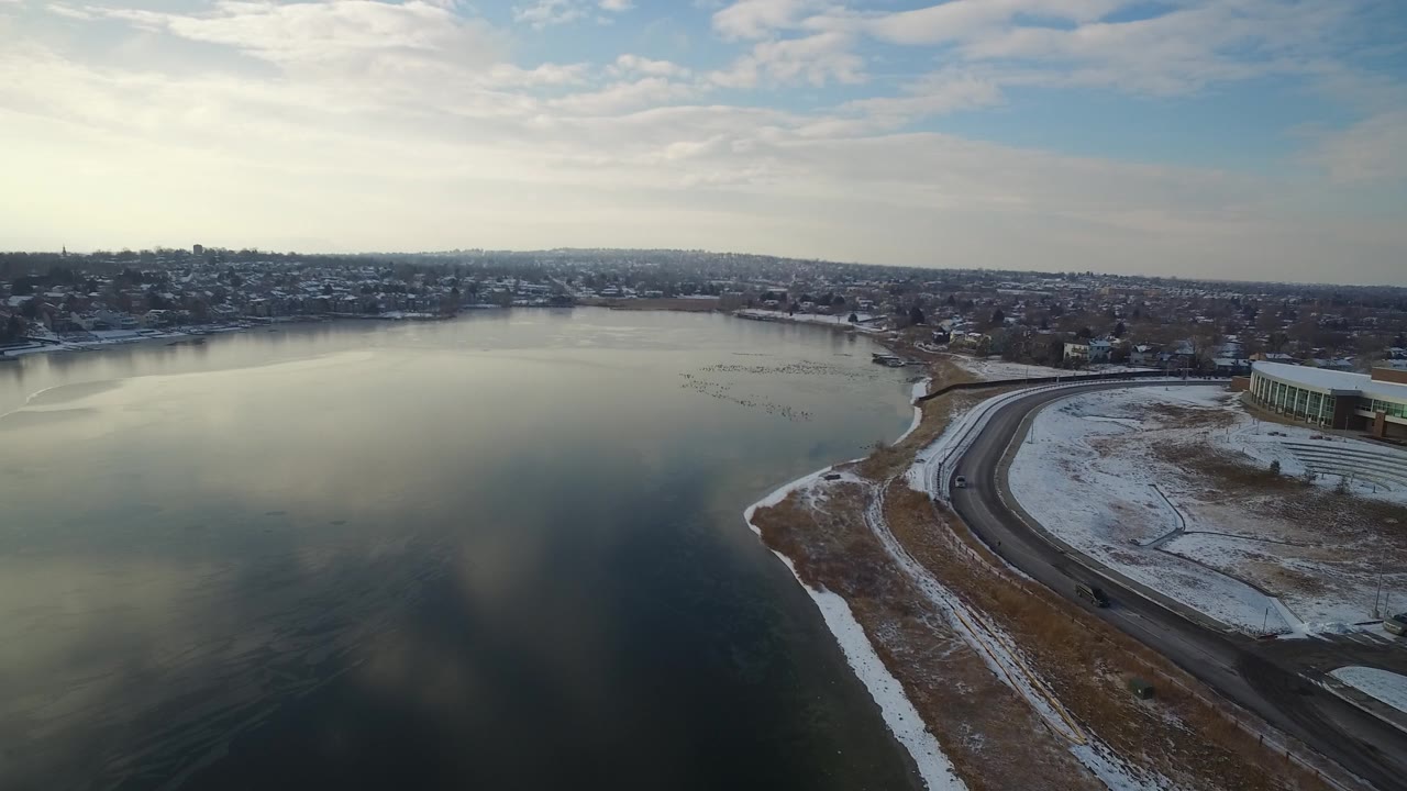 Aerial view at 0:01 over Hidden Lake, Colorado with the reflective lake filling most of the frame, the road bending along the snowy shore, a low lakeside building, and no visible people.
