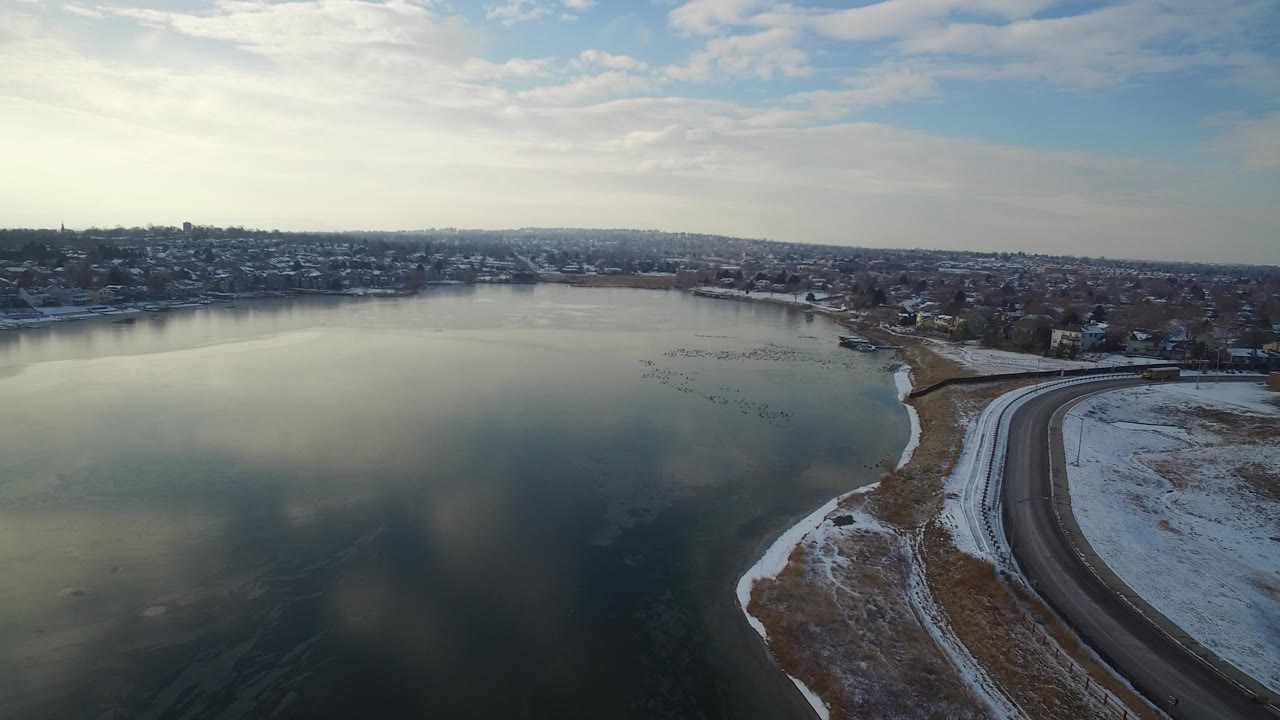 Frozen lake beside a snowy curved shoreline road and distant homes.