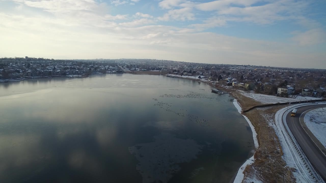 Wide opening aerial over a frozen lake in Hidden Lake, Colorado, with snowy shorelines, distant houses, a pale clouded sky, and no visible people.