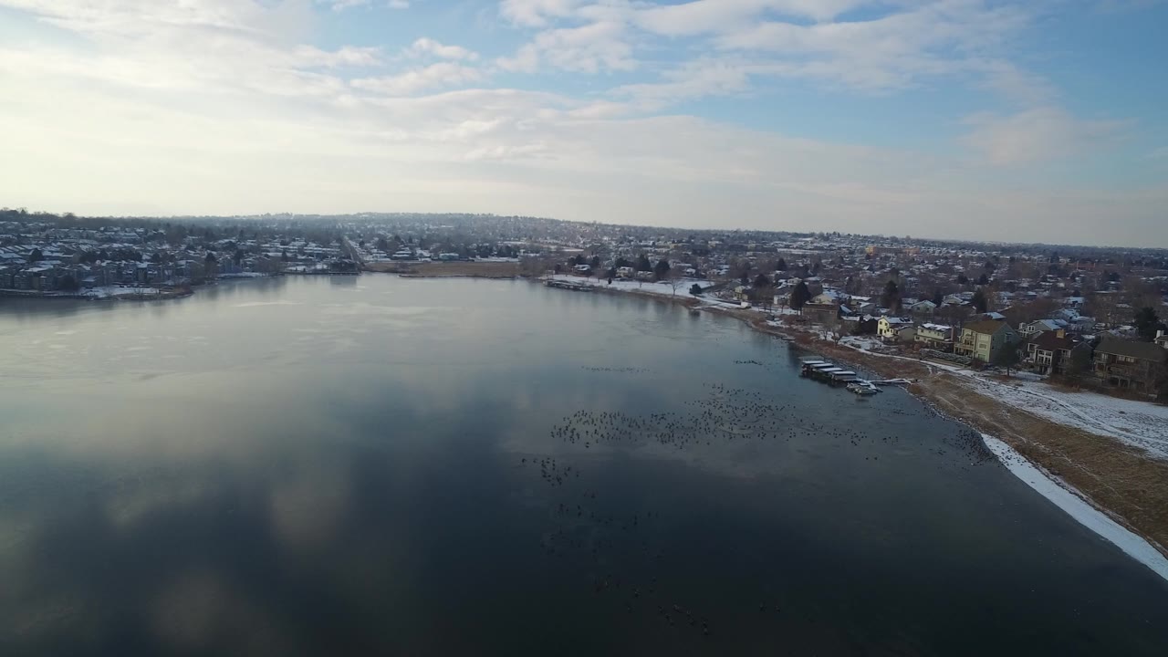 Aerial view over calm lake water and a snowy residential shoreline.