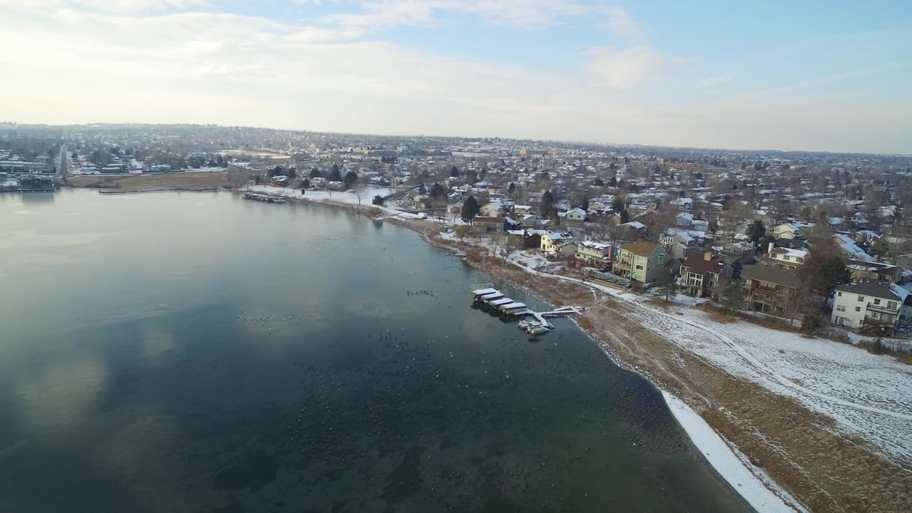 Aerial frame over Hidden Lake, Colorado showing dark lake water in the foreground, a snow-lined shore, small docks, lakeside houses, and no visible people.