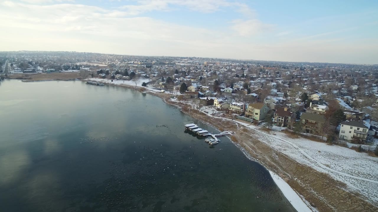 High aerial view of lakeside homes, docks, snow, and distant neighborhood blocks.