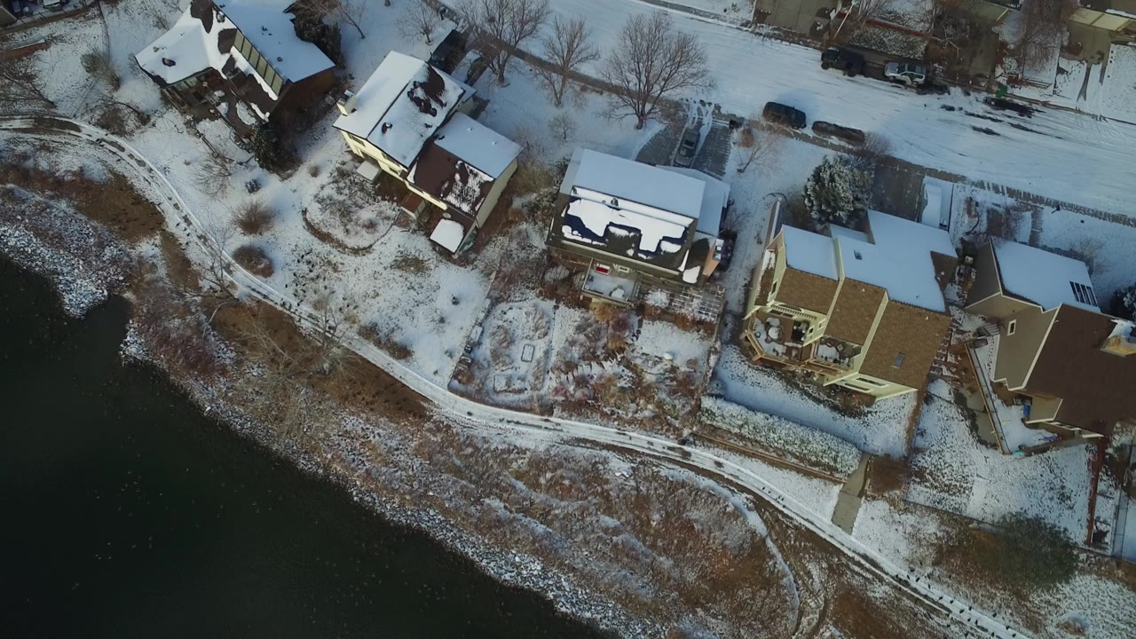 Overhead view in Hidden Lake, Colorado showing snow-covered homes beside dark water, a curved road, bare trees, and no visible people.