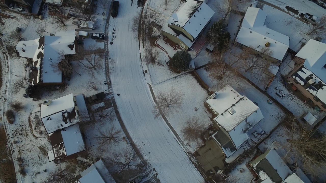 Overhead snowy neighborhood with houses, yards, bare trees, and a curved road.