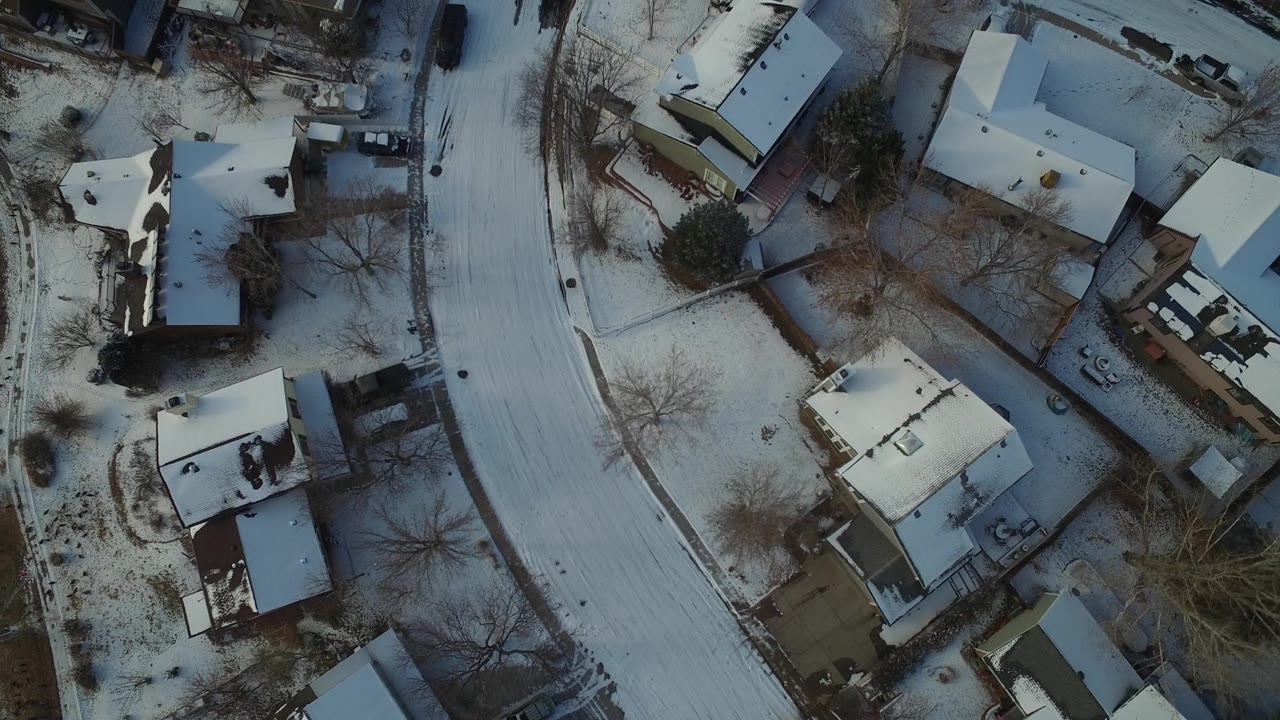 Near-vertical view of Hidden Lake, Colorado rooftops and yards beside a pale snow-covered road, with leafless trees casting winter shadows and no visible people.