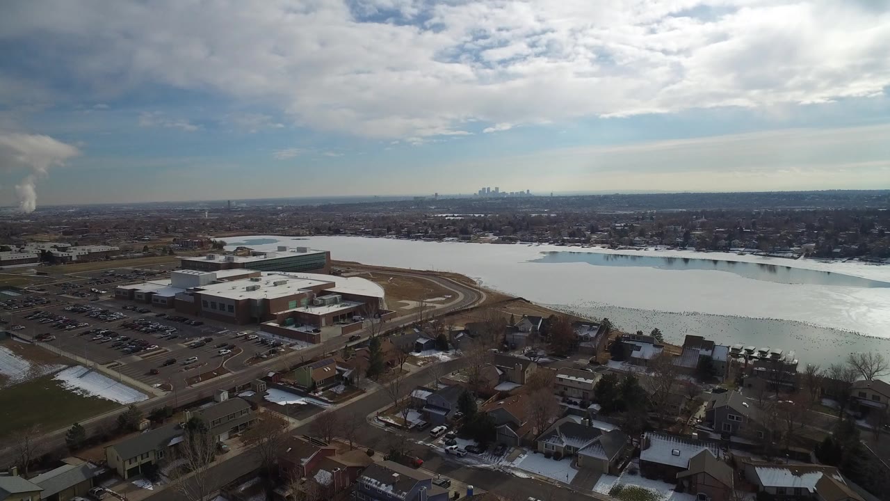 Aerial frame from Hidden Lake, Colorado, showing a large flat-roofed building beside a broad waterway, homes in the foreground, distant skyline on the horizon, and no clearly visible people.