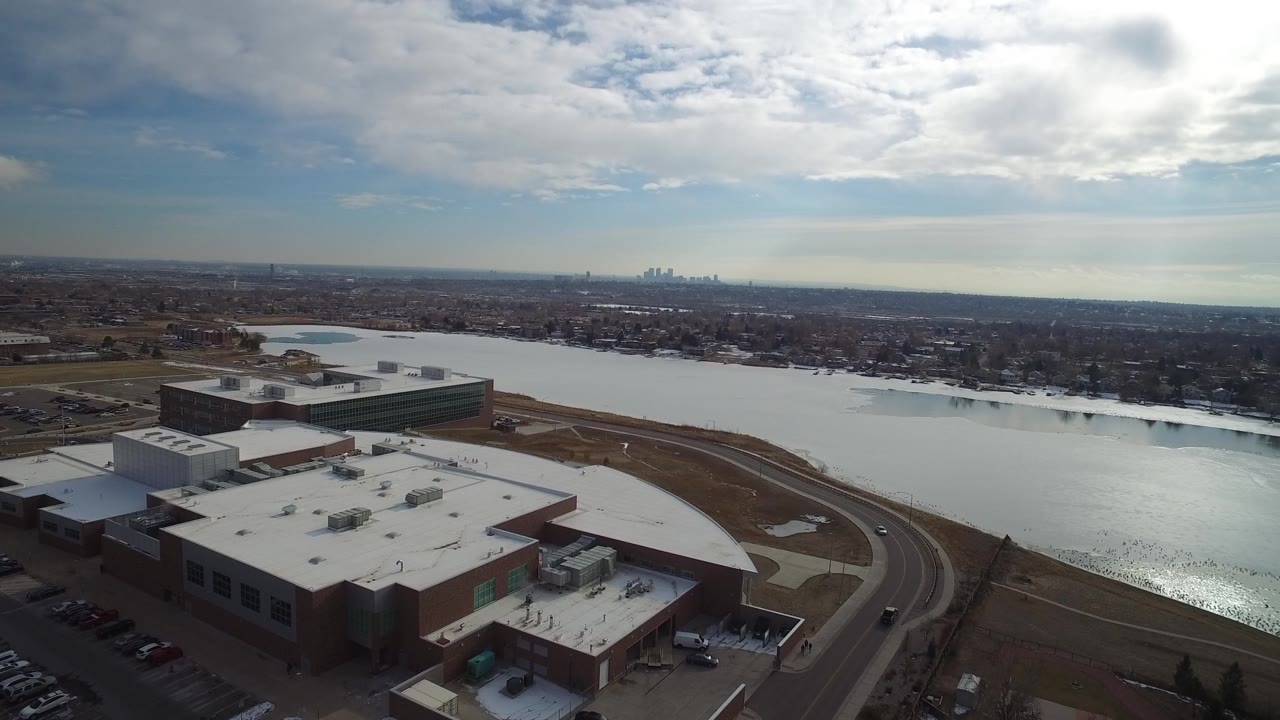 Aerial view of large flat-roofed buildings beside a wide lake under cloudy sky.