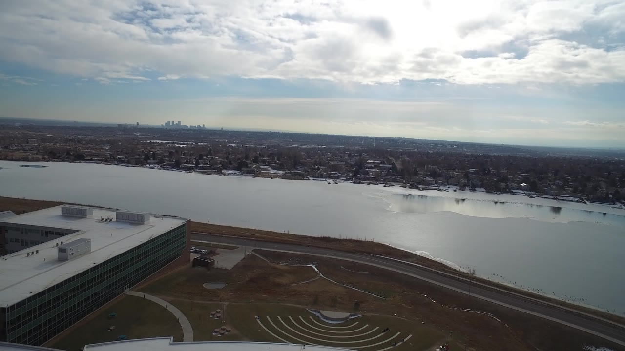 Wide aerial over lake water with a large building roof at bottom and skyline in the distance.