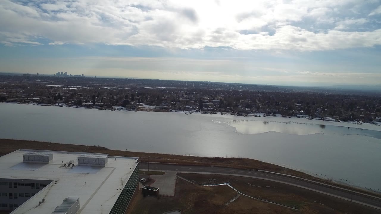 Aerial view of a lake, shoreline, rooftop, distant skyline, and clouded sky.