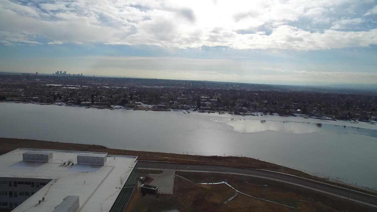 Aerial view over Hidden Lake, Colorado with the rooftop shifted across the lower foreground, pale water reflecting the sky, low buildings beyond the shore, and no visible people.