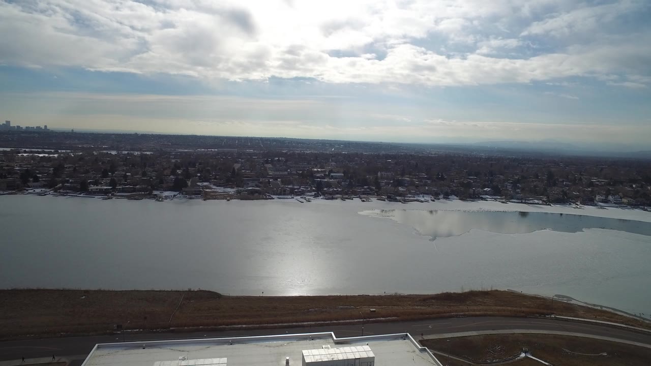 Opening aerial frame over Hidden Lake, Colorado, showing a frozen lake with sun glare, a brown shoreline road in front, town blocks across the water, and no visible people.