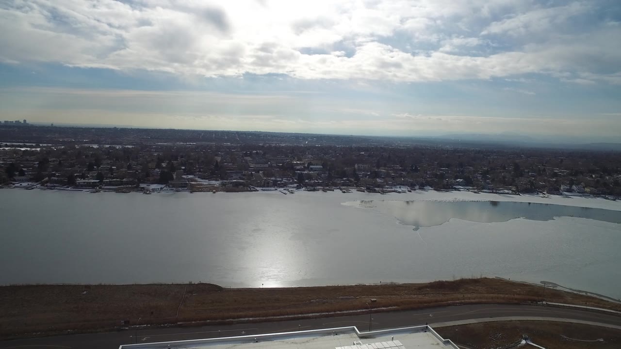Wide drone frame of frozen lake with broken cloud light over a distant town.