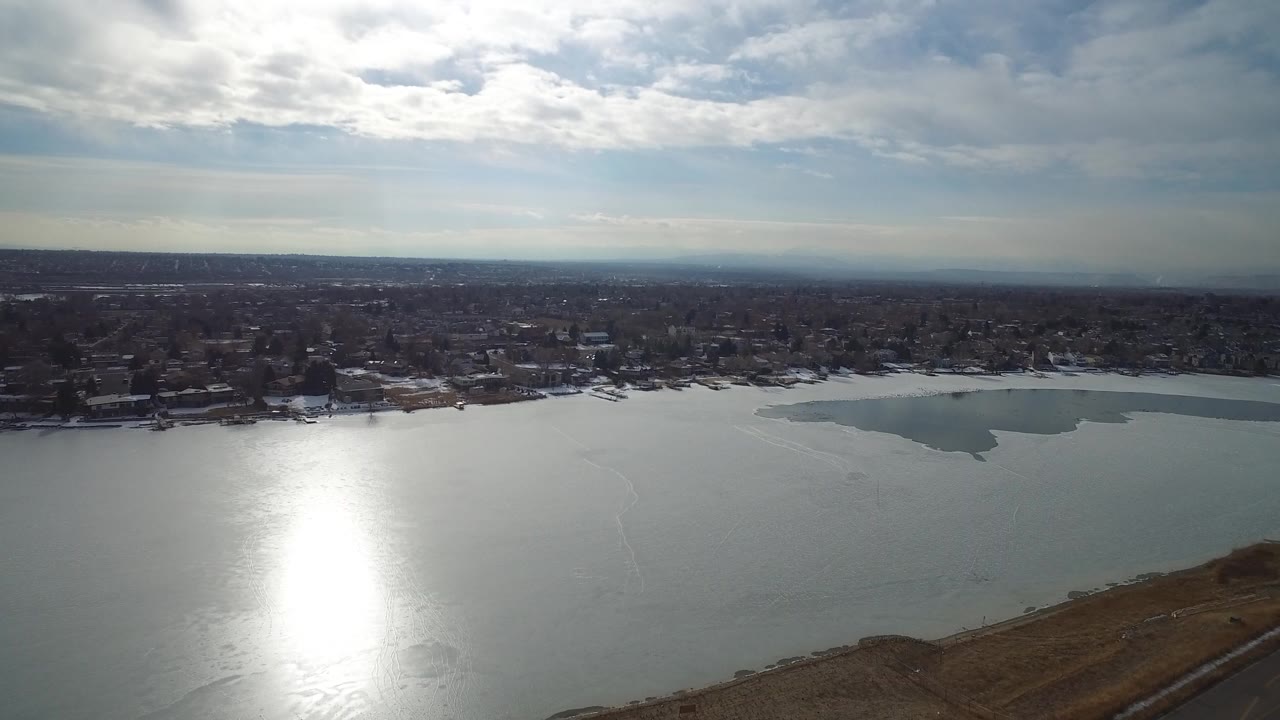 Aerial view over a lake with sun glare, shoreline homes, and cloudy sky.