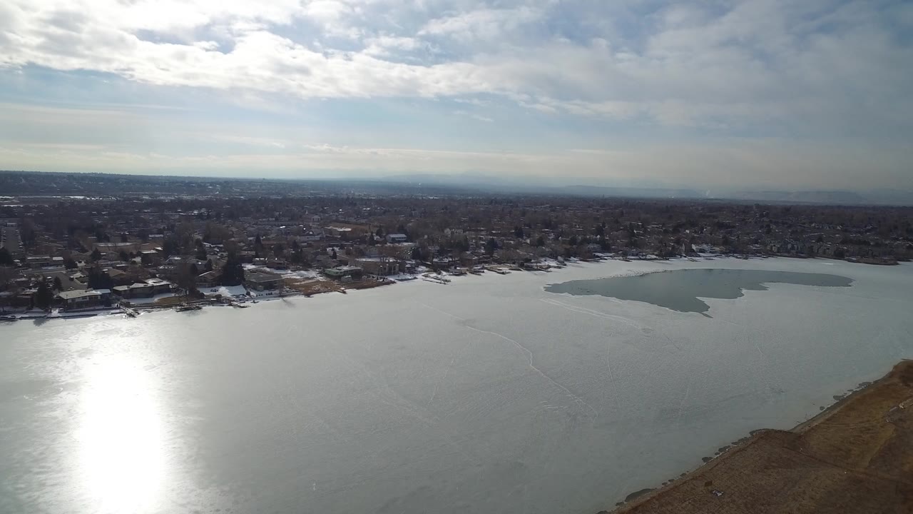 Opening aerial frame over Hidden Lake, Colorado showing reflective water, distant shoreline homes, a pale patch on the lake, brown foreground land, and no visible people.