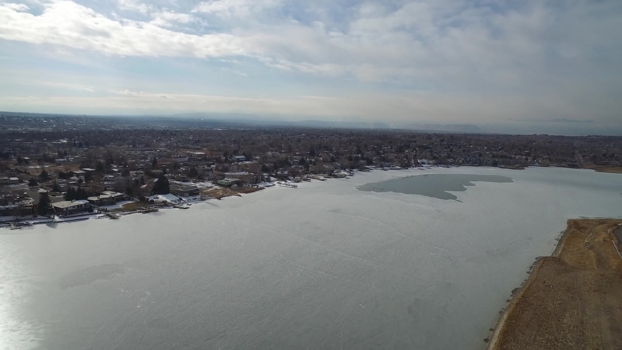 Drone-height view across a lake toward homes with a pale ice-like patch near the far shore.