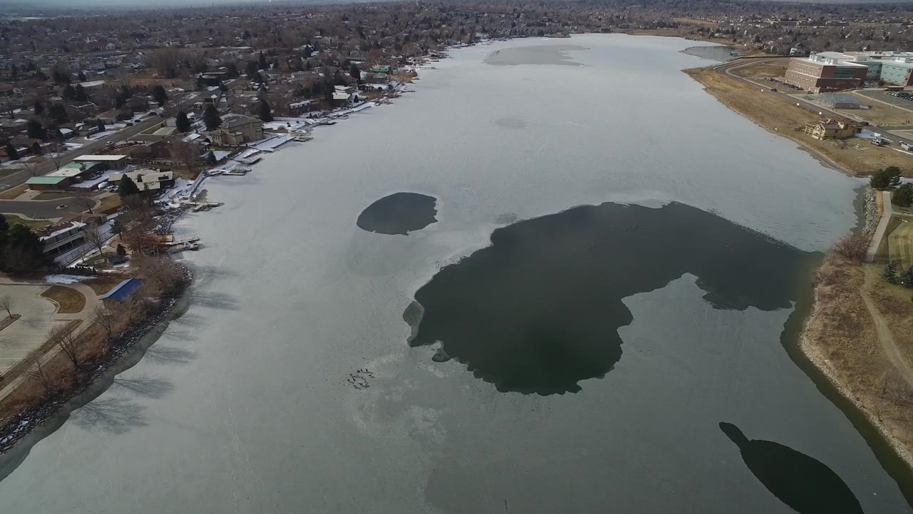 Aerial view over Hidden Lake, Colorado, with a large dark opening in the ice, smaller open patch farther back, houses along the left shore, commercial buildings at upper right, and no visible people.