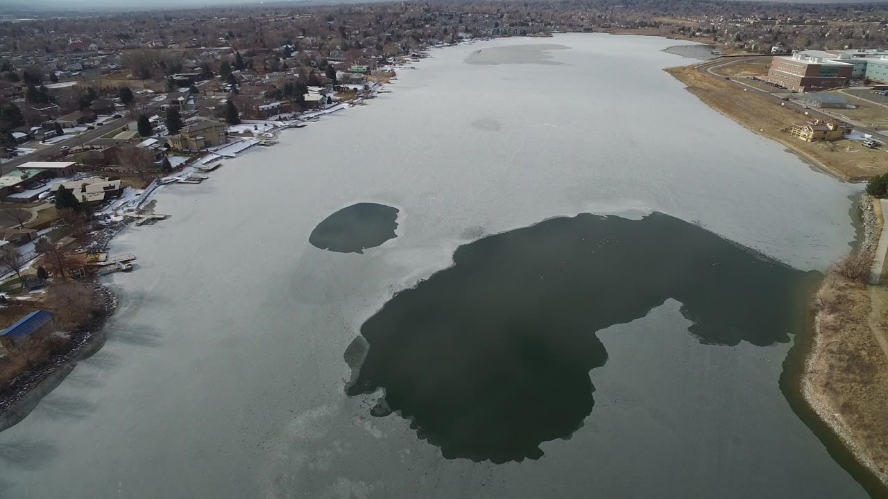 Aerial view of a partly frozen lake bordered by homes and open land.