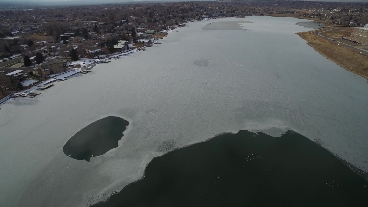 High aerial view over Hidden Lake, Colorado, with pale ice, dark open-water patches, shoreline houses, docks, snow along the bank, and no visible people.