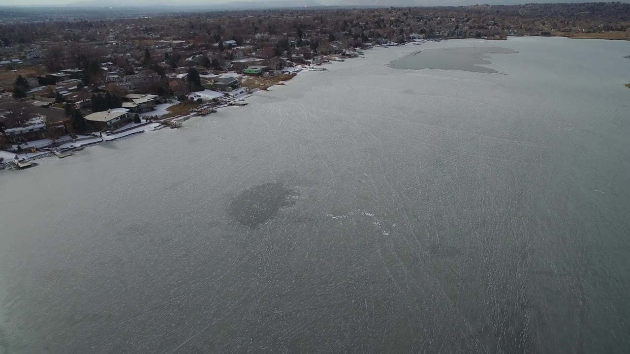 Aerial frame looking across rippled Hidden Lake water toward a row of houses, docks, and winter trees, with no visible people.