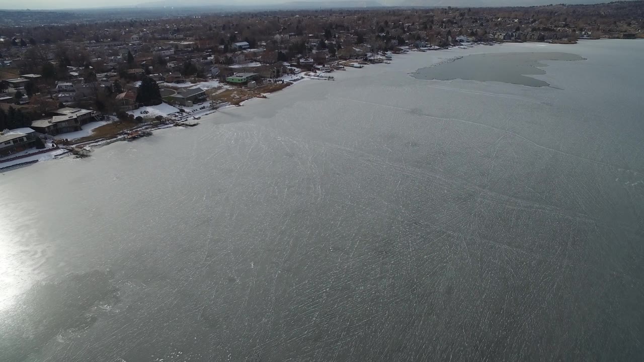 Aerial frame over Hidden Lake, Colorado, showing bright glare on the icy surface, shoreline buildings at the top of frame, and no visible people.