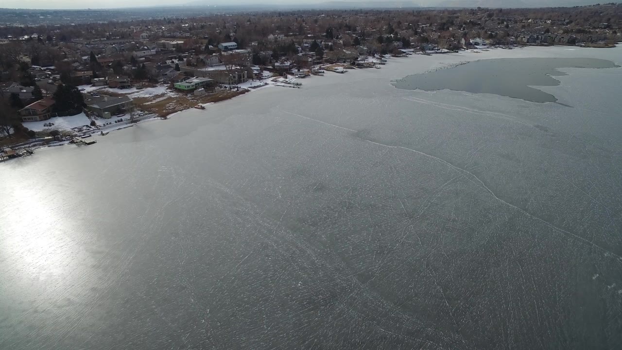 Aerial view over a frozen lake with shoreline homes and sun glare on the ice.