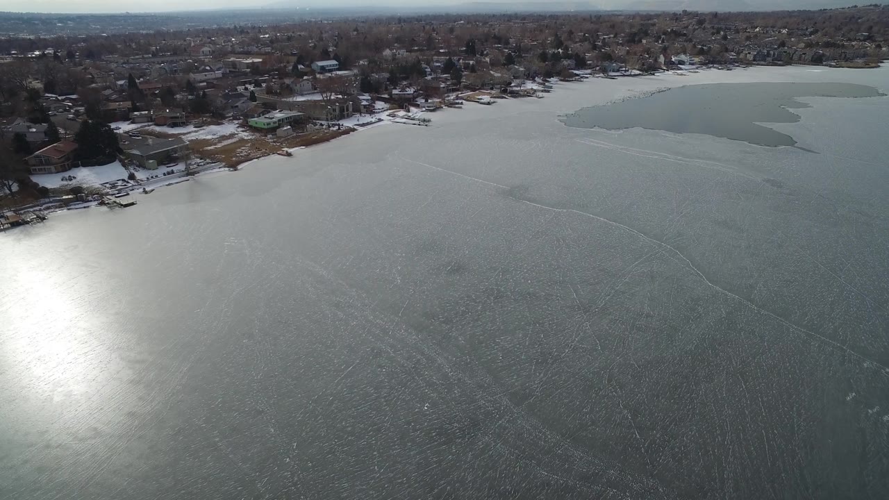 Elevated view over Hidden Lake, Colorado, showing textured gray ice, a bright reflection near the lower left, waterfront homes along the far shore, and no visible people.