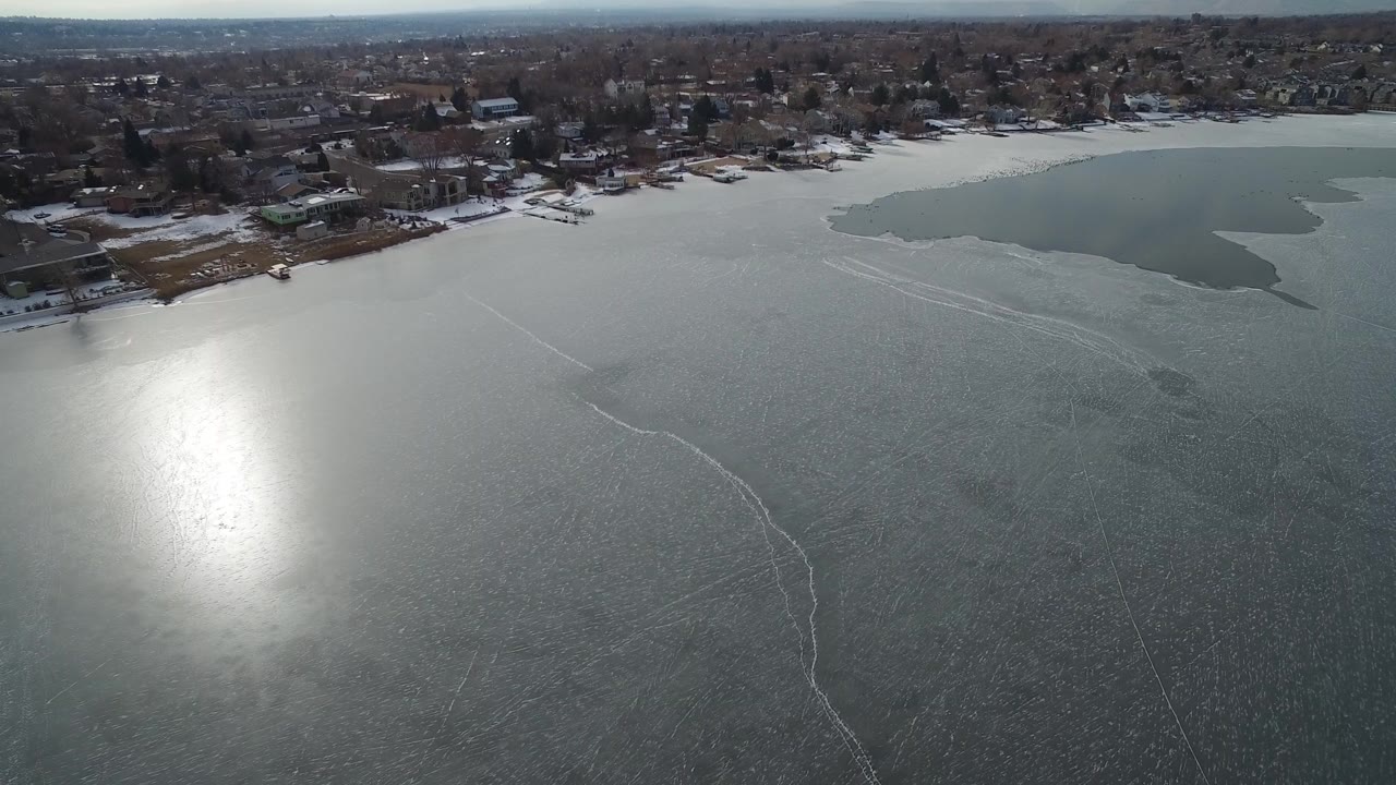 Aerial view over a partly frozen lake with shoreline homes and sun glare.