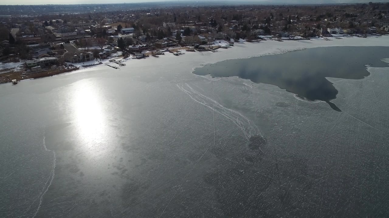 Aerial view of icy lake with bright sun reflection and homes along the far shore