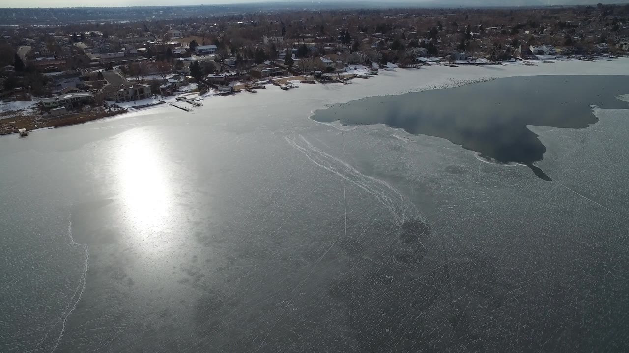 Aerial view of Hidden Lake, Colorado with sun glare shifted across gray ice, faint white tracks on the lake surface, dark open water near the upper right, homes on the shore, and no visible people.