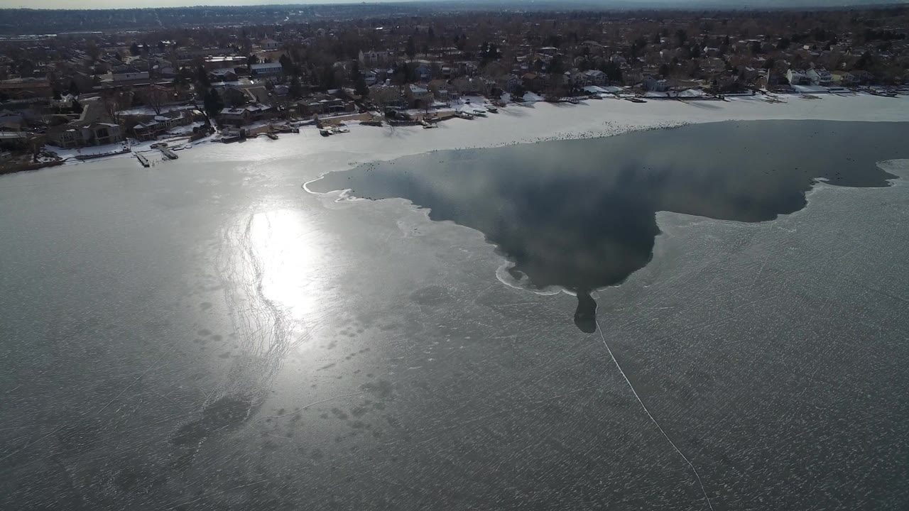 Aerial view of a partly frozen lake with bright sun glare and shoreline homes.
