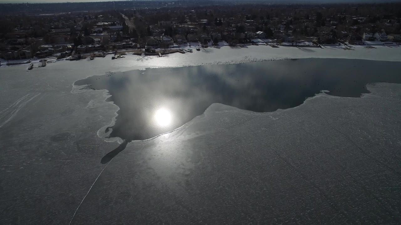 Aerial view of a partly frozen lake with sun glare and shoreline homes.