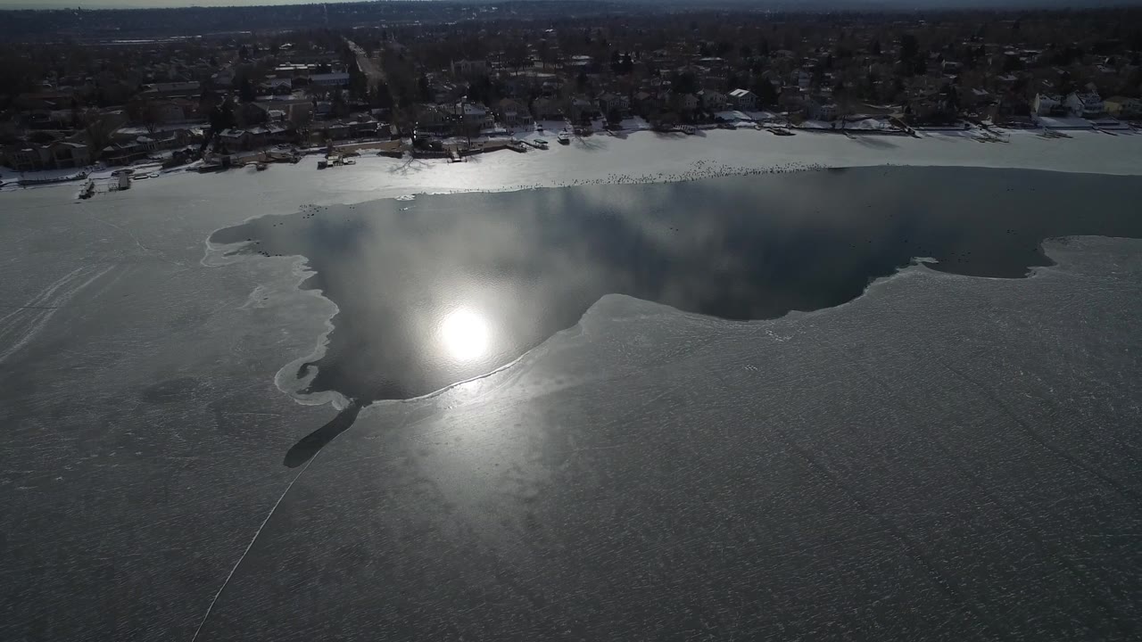 Aerial frame over Hidden Lake, Colorado, with the sun reflection shifted across the ice, open water stretching along the far bank, shoreline trees, distant homes, and no visible people.