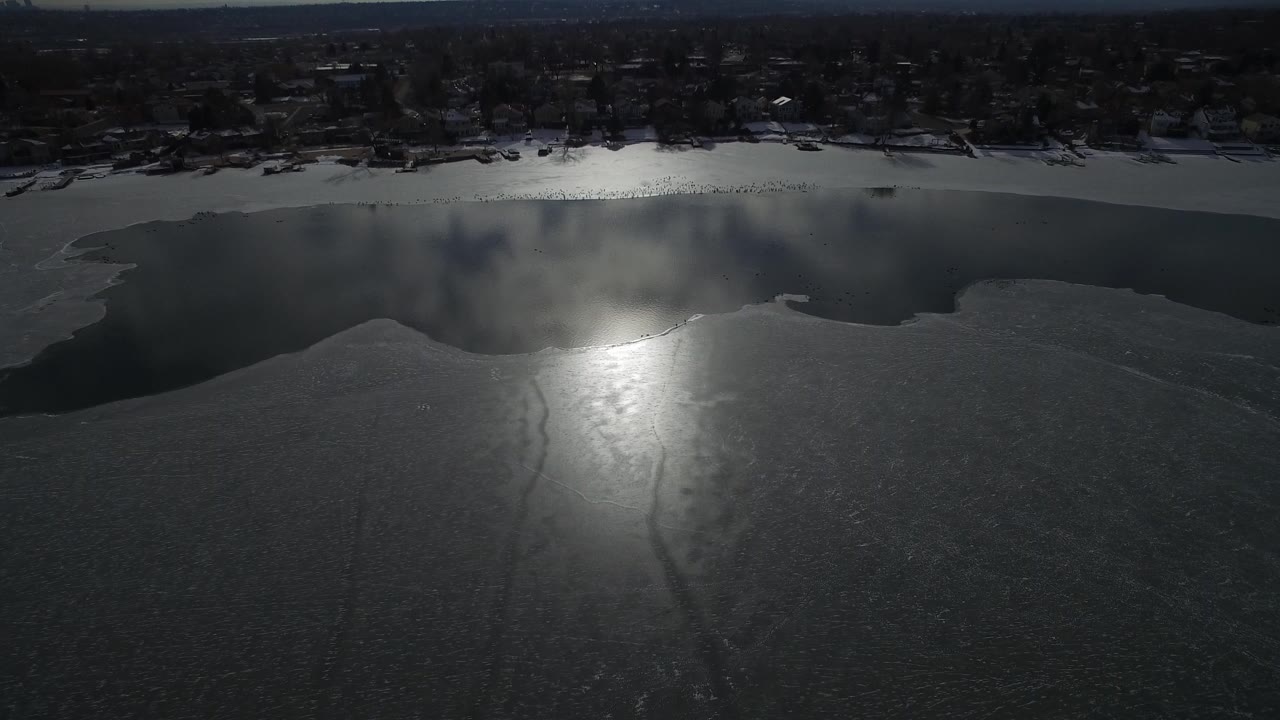 Aerial frame over Hidden Lake, Colorado showing dark ice, a bright sun reflection near center, open water by the far shore, distant homes, and no visible people.