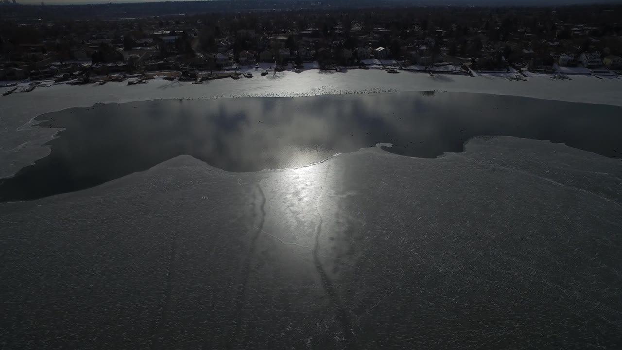 Hidden Lake, Colorado aerial view with the sun reflection shifted across the ice, open water along the upper shoreline, winter trees, distant houses, and no visible people.