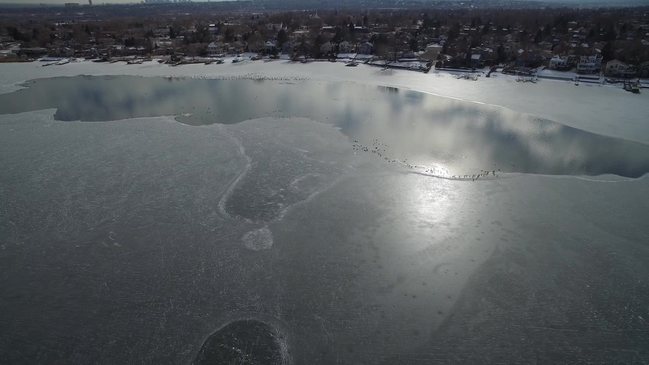 Aerial frame over Hidden Lake, Colorado showing gray ice, darker open water near the snowy shoreline, distant homes and bare trees, and no visible people.