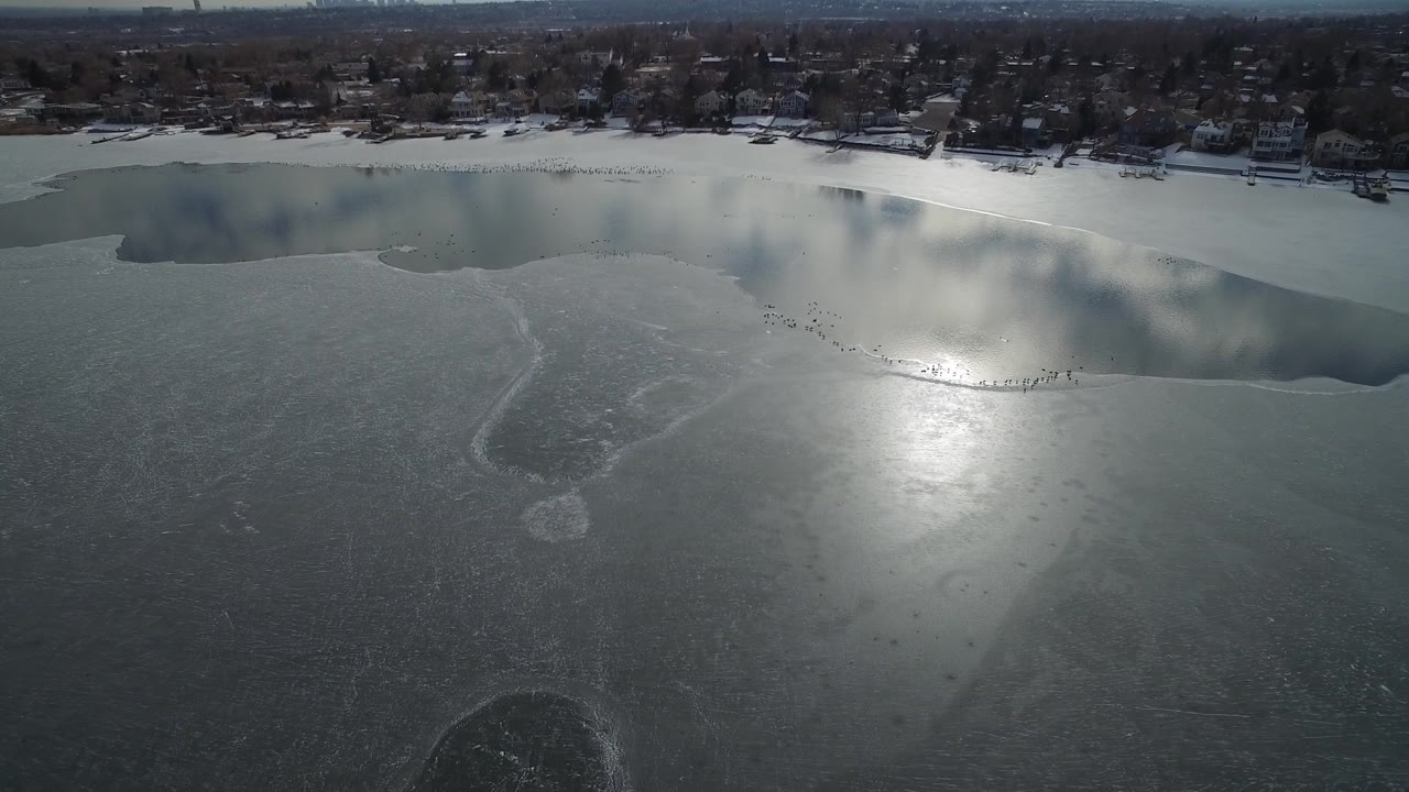 Hidden Lake, Colorado appears from above with a bright reflection on the ice, an irregular open-water edge near the top of frame, shoreline homes, and no visible people.