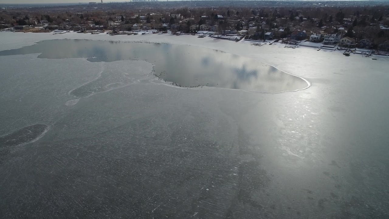 Aerial frame over Hidden Lake, Colorado showing a wide gray ice surface, bright sun reflection near center, shoreline houses and leafless trees in the distance, with no visible people.
