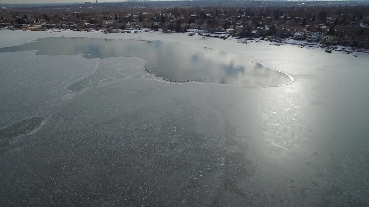 Aerial view in Hidden Lake, Colorado with a curved lighter ice boundary across the water, a bright reflective patch on the lake, distant homes, winter trees, and no visible people.