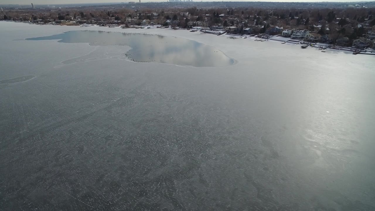 Aerial frame from Hidden Lake, Colorado showing broad gray ice, a brighter reflection across the lake, darker water near the upper shoreline, and no visible people.