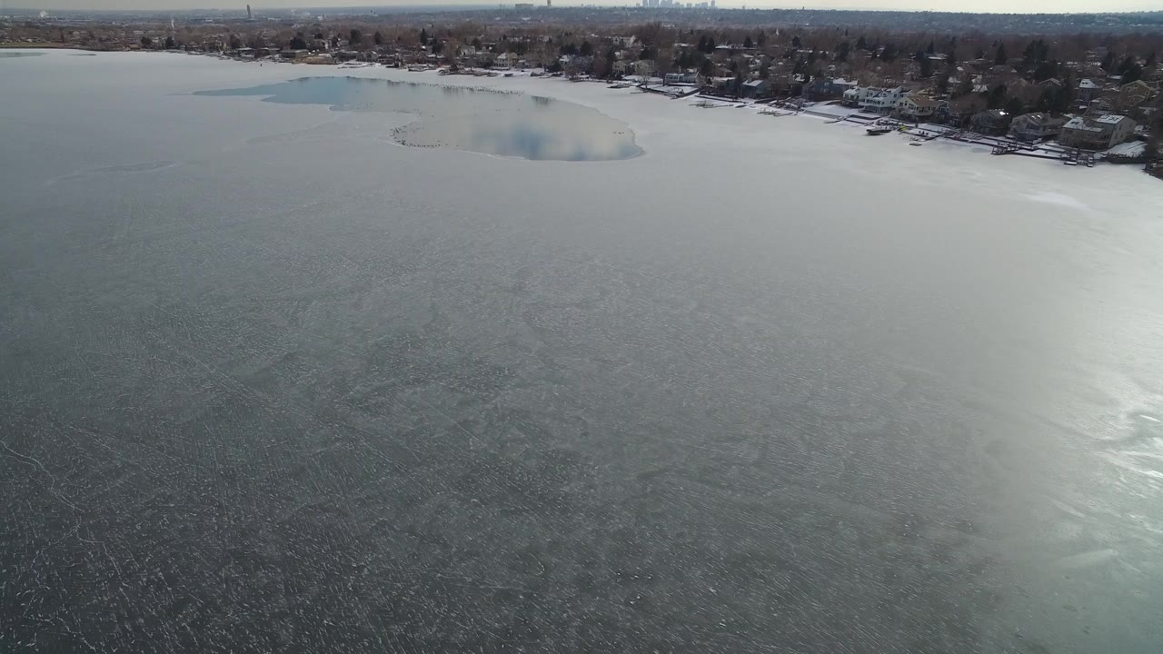 Aerial view over a gray partially frozen lake with shoreline homes beyond.
