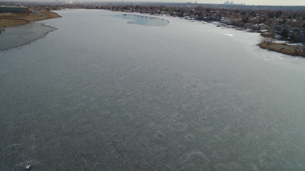 Drone frame over the frozen Hidden Lake water, showing a curved residential shoreline, dark trees, light snow patches, and no visible people.