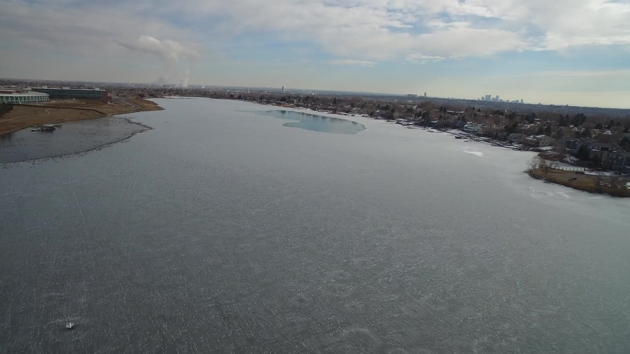 Aerial view over a broad gray lake with shoreline homes and distant buildings.