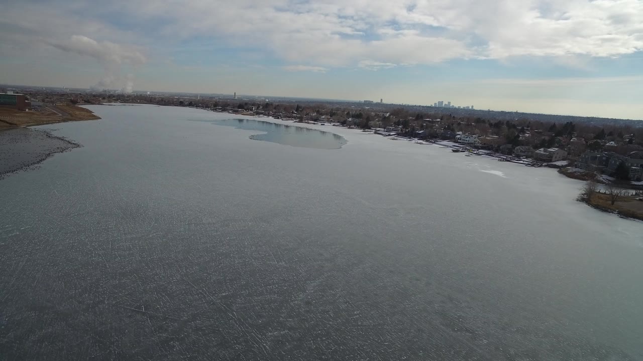 Opening aerial frame over Hidden Lake, Colorado, showing textured gray water in the foreground, houses along the right shore, brown land at left, distant buildings on the horizon, and no visible people.
