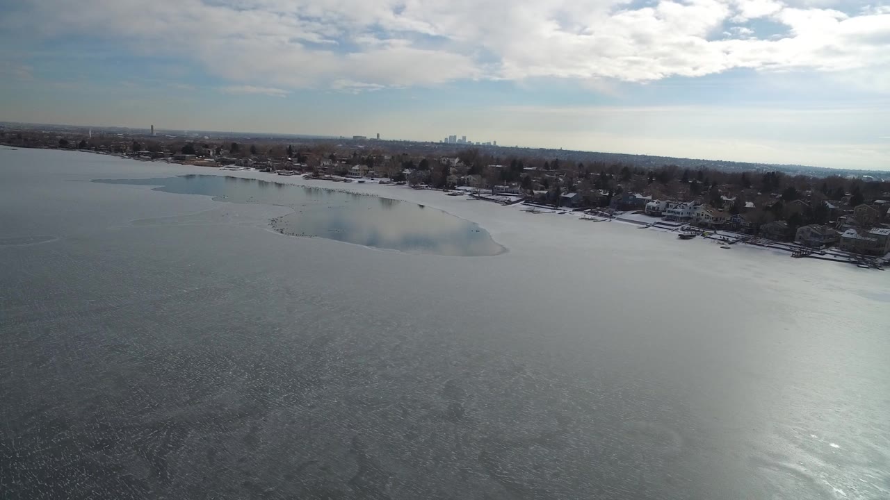 Aerial view over a frozen lake with a snowy residential shoreline.