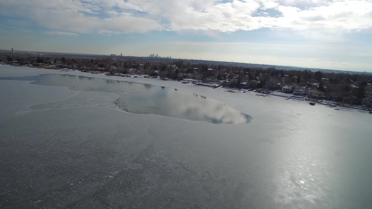 Aerial frame over Hidden Lake, Colorado, showing gray frozen water in the foreground, shoreline houses and trees beyond it, a distant skyline, bright cloud cover, and no visible people.