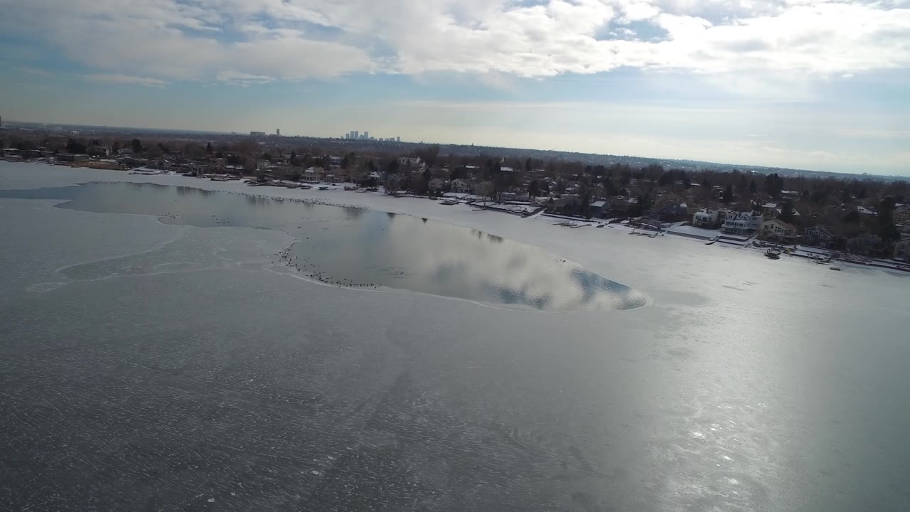 Wide aerial lake view with frozen surface, shoreline neighborhood, and skyline on the horizon.