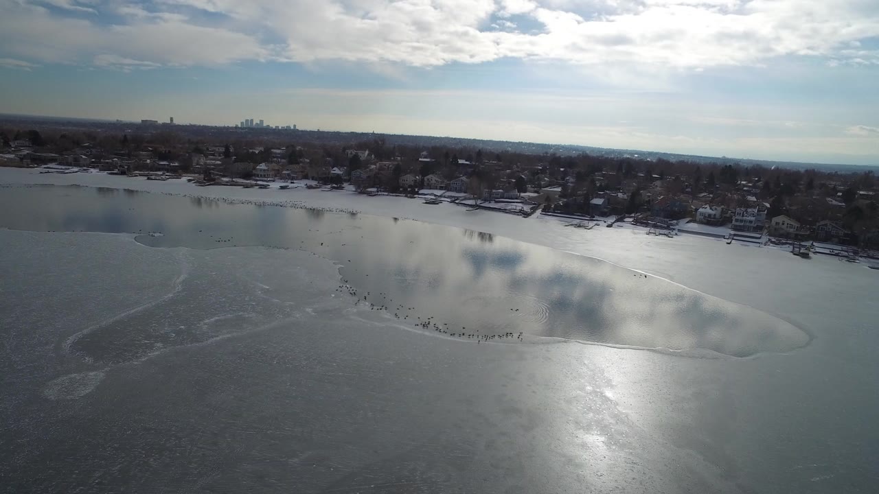 High aerial view over a partly frozen lake and distant shoreline homes.