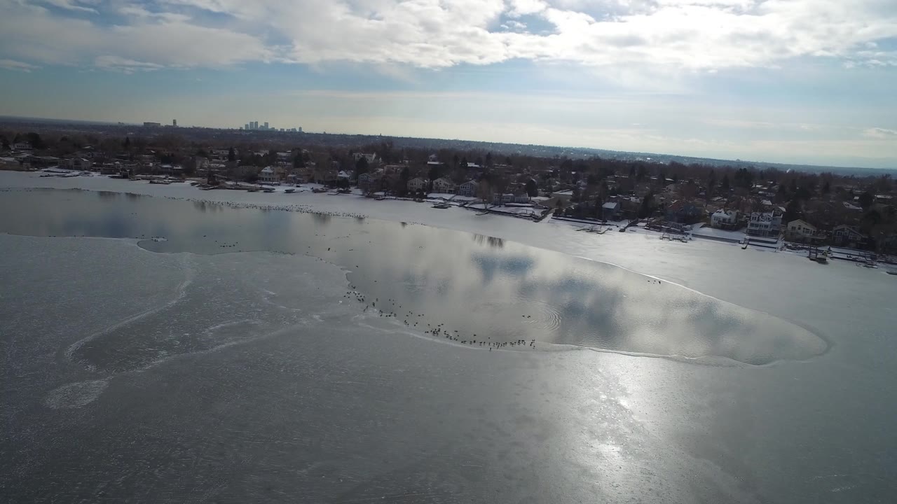 Aerial view of Hidden Lake in Colorado showing a broad frozen surface, a bright reflection near center, shoreline houses, cloud shadows, and no visible people.