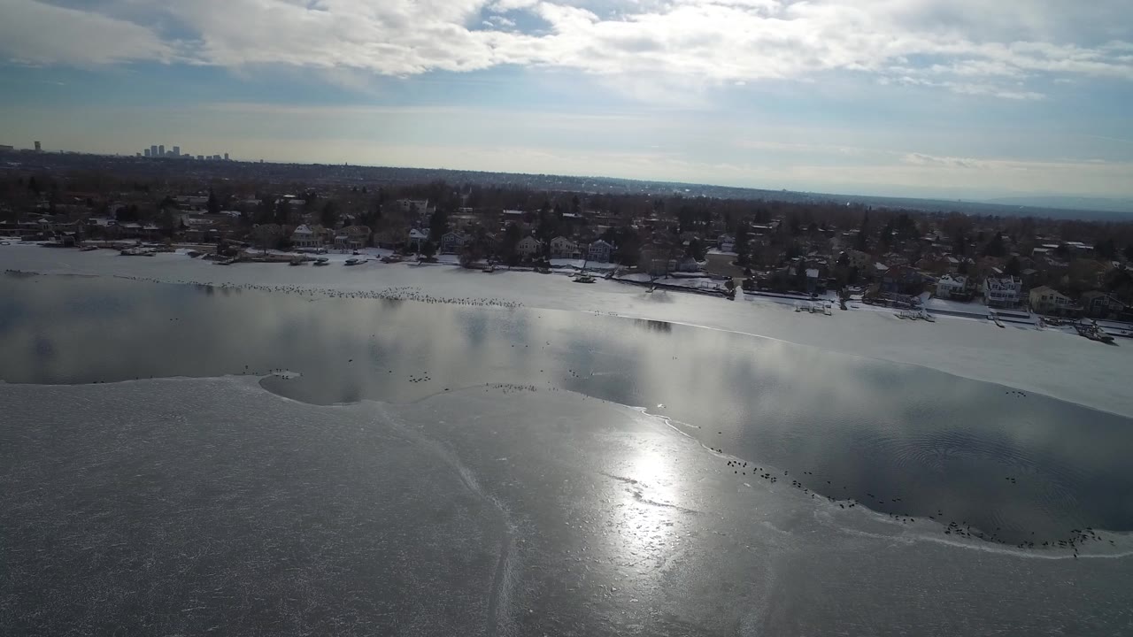 Aerial view over Hidden Lake, Colorado, with dark water, pale ice in the foreground, houses and trees along the shore, a distant skyline, and no visible people.