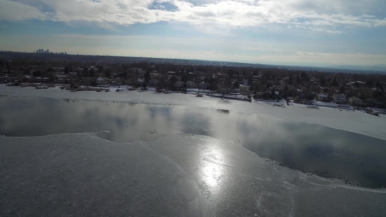 Winter aerial over ice and open water with a residential shore in the distance.